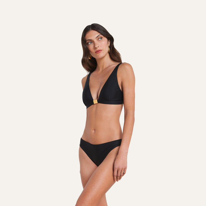 Wearing the Nicole Bikini Bottom, a woman stands against a plain light background. The black bikini bottom, crafted from four-way stretch nylon for comfort and featuring moderate coverage with gold accents, is paired with a matching bikini top as she looks to the side.
