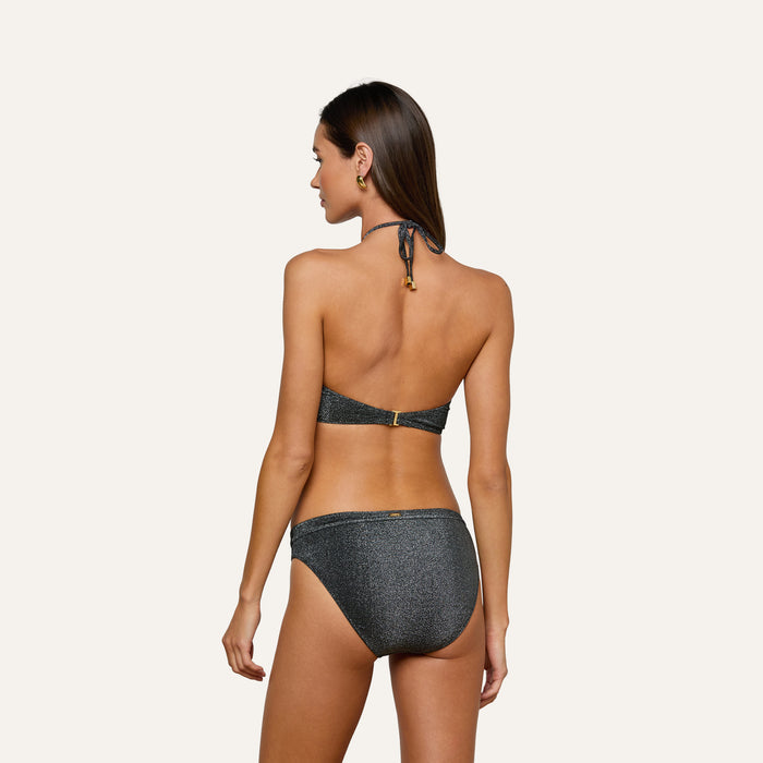 Wearing the Nicole Bikini Bottom in metallic black and gold hoop earrings, a woman with long brown hair stands against a plain light background, shown from the back.