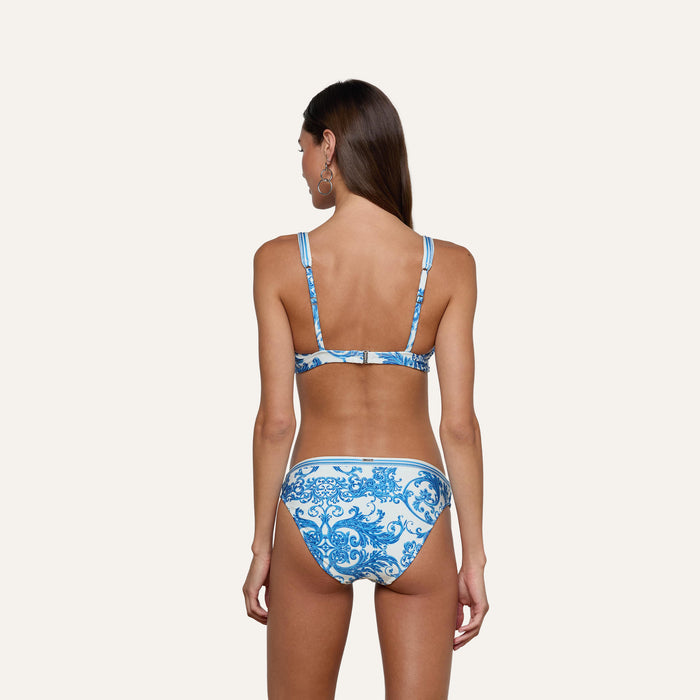 A woman with long brown hair, seen from behind, models the Nicole Bikini Bottom in a white and blue baroque print featuring moderate coverage and adjustable straps, paired with hoop earrings, against a plain light background.