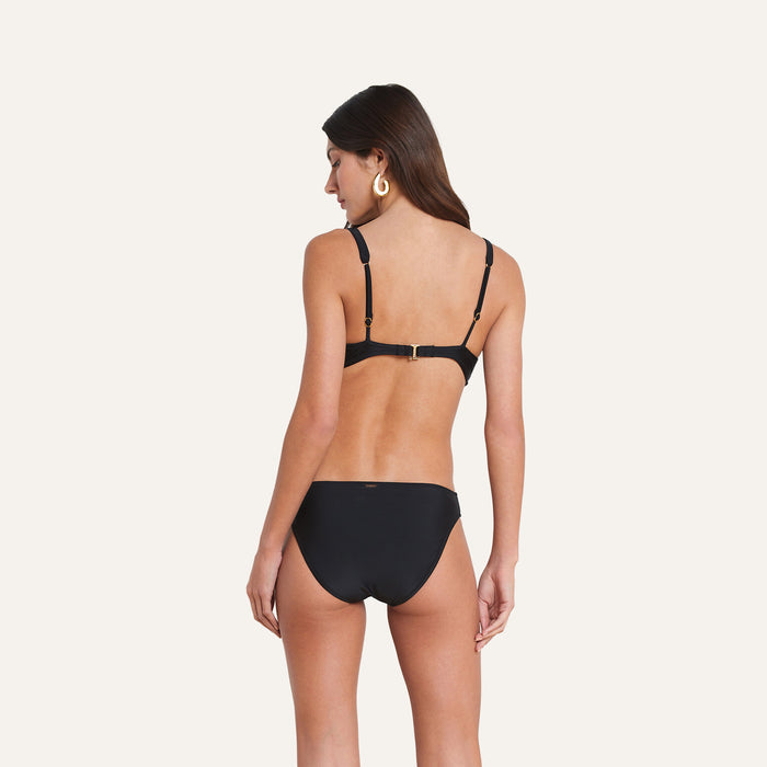 A woman with long brown hair is shown standing with her back to the camera against a plain light background, wearing the Nicole Bikini Bottom in black, crafted from four-way stretch nylon for moderate coverage, paired with large hoop earrings.