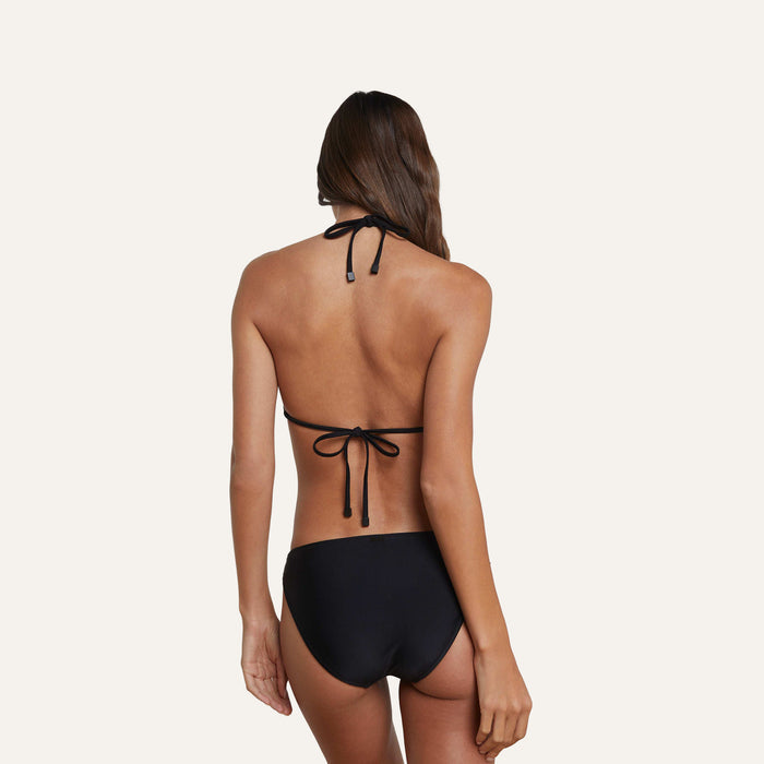 A woman with long brown hair is shown from the back, wearing versatile black halter-neck swimwear paired with the Nicole Bikini Bottom, which offers moderate coverage, against a plain light background.