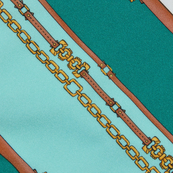 Nicole Bikini Bottom in a patterned fabric with teal and light blue stripes, featuring a belt-stripe print accented by brown leather straps and gold chains running diagonally across the surface.