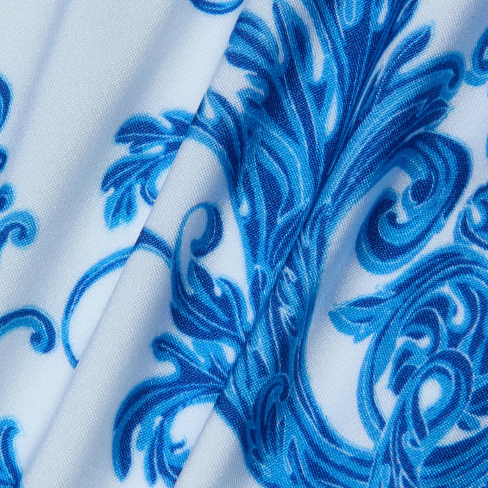 Introducing the Nicole Bikini Bottom, featuring white fabric adorned with an intricate blue baroque print of floral and swirl motifs. Slightly folded to reveal texture and detail, this piece is crafted from four-way stretch nylon for moderate coverage.