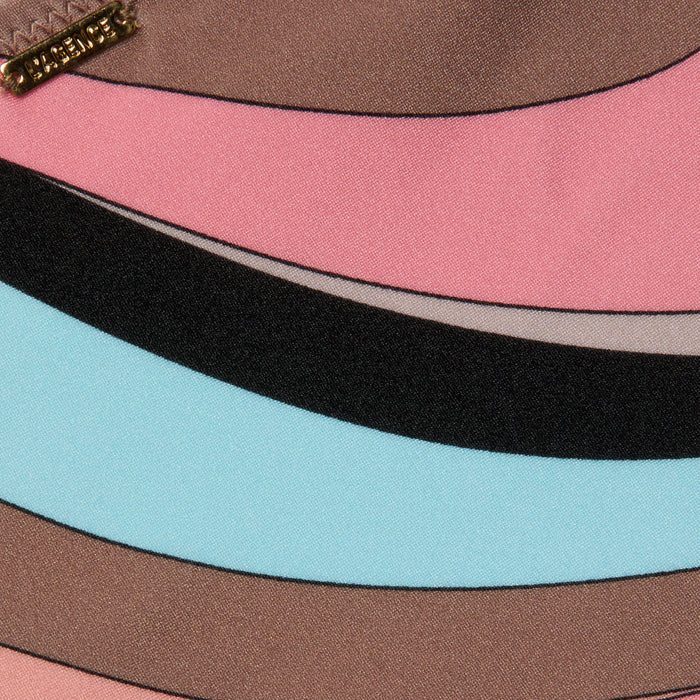 Close-up of the Nicole Bikini Bottom fabric with wavy stripes in pink, brown, black, and light blue, featuring an abstract swirl print. A small metallic tag with engraved text is sewn into the upper left corner.