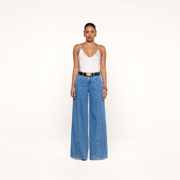 A person stands against a white background wearing a white camisole top, the Niles Wide-Leg Jean in a high rise lightweight denim, a black belt with a large gold buckle, and hoop earrings.