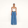 A person stands against a white background wearing a white camisole top, the Niles Wide-Leg Jean in a high rise lightweight denim, a black belt with a large gold buckle, and hoop earrings.