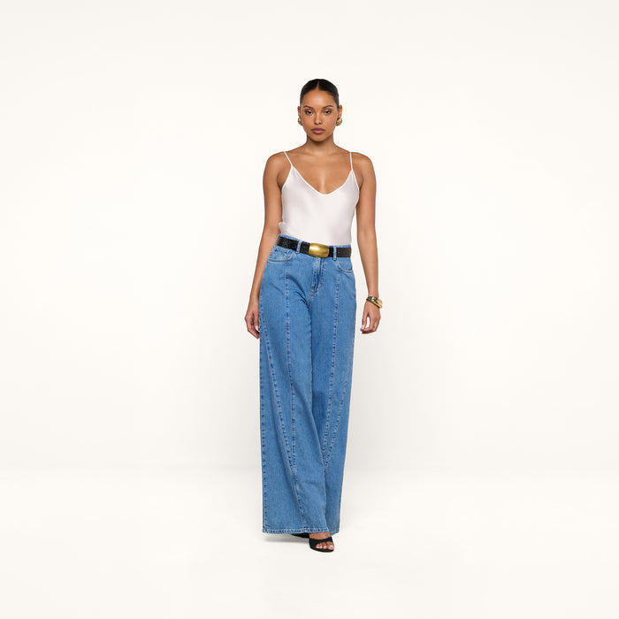A woman stands against a plain white background wearing a white tank top, the Niles Wide-Leg Jean styled with a black belt, black open-toe heels, and gold jewelry.