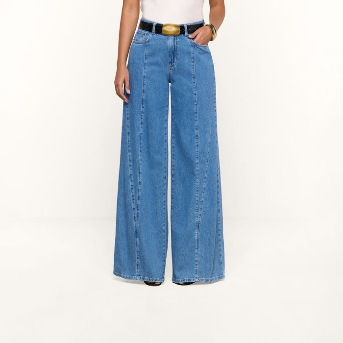 A person wearing the Niles Wide-Leg Jean, featuring a high-rise, wide-leg silhouette in lightweight blue denim, styled with a black belt and white top, stands against a plain white background.