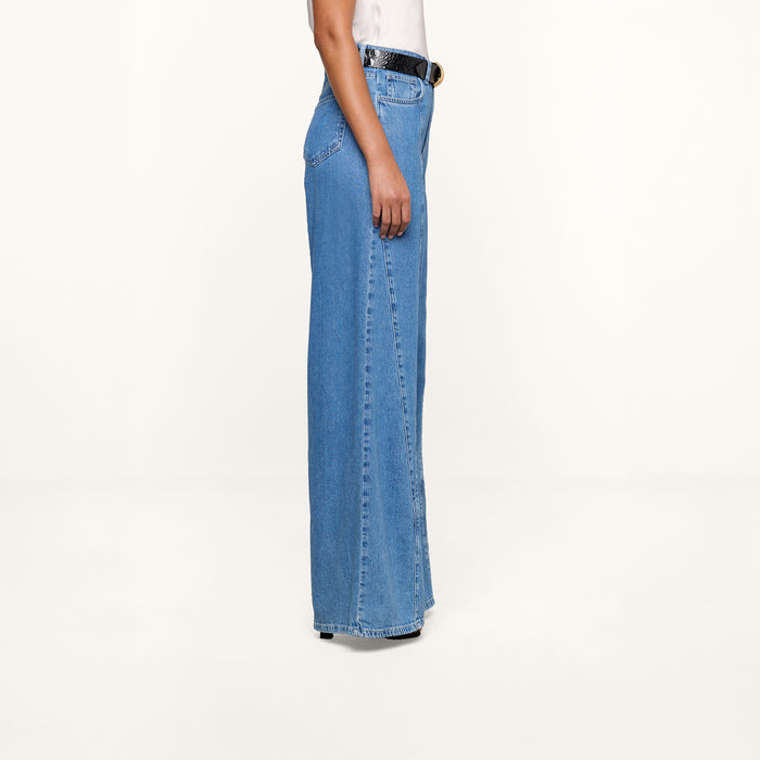 Sure Here’s the revised sentence using your product name: A person stands sideways against a plain white background, wearing a white top tucked into high rise Niles Wide-Leg Jeans made from lightweight blue denim, styled with a black belt.