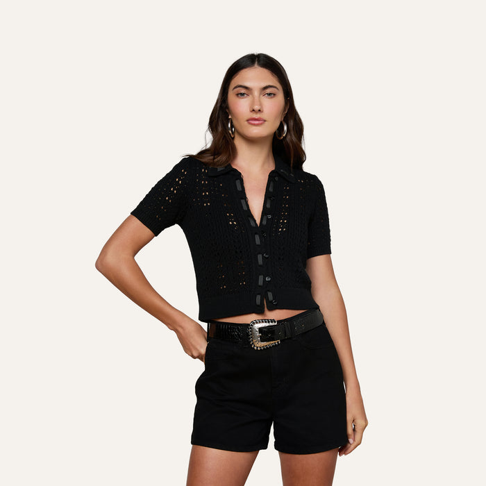 A woman stands against a light background, wearing the Ninette Knit Top—a black short-sleeve pointelle knit top with buttons—paired with black shorts, a black belt featuring a decorative buckle, and hoop earrings. One hand rests on her hip, exuding Parisian flair.