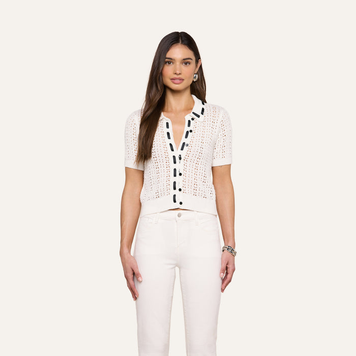 A woman with long brown hair wears the Ninette Knit Top, a short-sleeve white shirt with black button trim, paired with white pants. She stands facing forward against a plain, light background.
