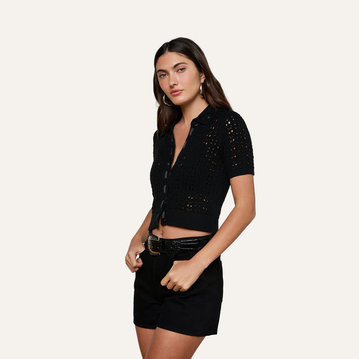 Wearing the Ninette Knit Top—a black short-sleeve, button-up crochet design with grosgrain ribbon trim—a woman poses with black belted shorts against a plain, light background.