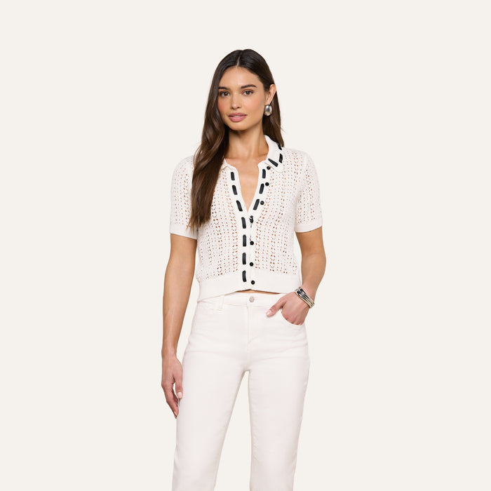 A woman stands against a plain background, wearing the Ninette Knit Top—a white short-sleeve, button-up pointelle knit cardigan with black grosgrain ribbon trim—paired with high-waisted white pants. Her right hand rests in her pocket, and she has long brown hair.