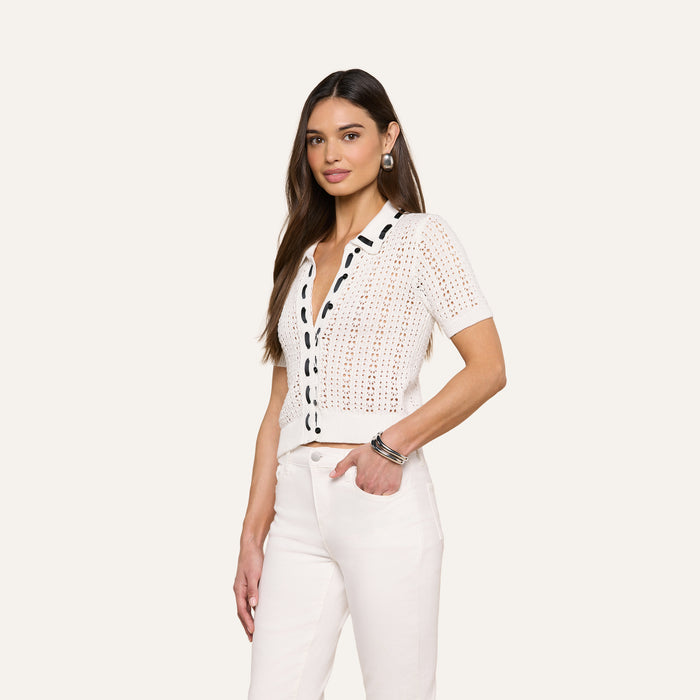 A woman with long brown hair is wearing the Ninette Knit Top—a white, short-sleeved top with black trim and Parisian flair—paired with white pants, standing with one hand in her pocket against a plain light background.