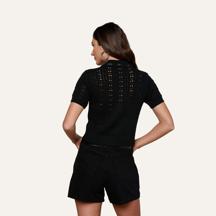 A woman with long brown hair stands facing away, wearing the Ninette Knit Top—a short-sleeve black pointelle knit top—paired with black shorts. Her hands rest on her hips, and she wears large hoop earrings against a plain light background.