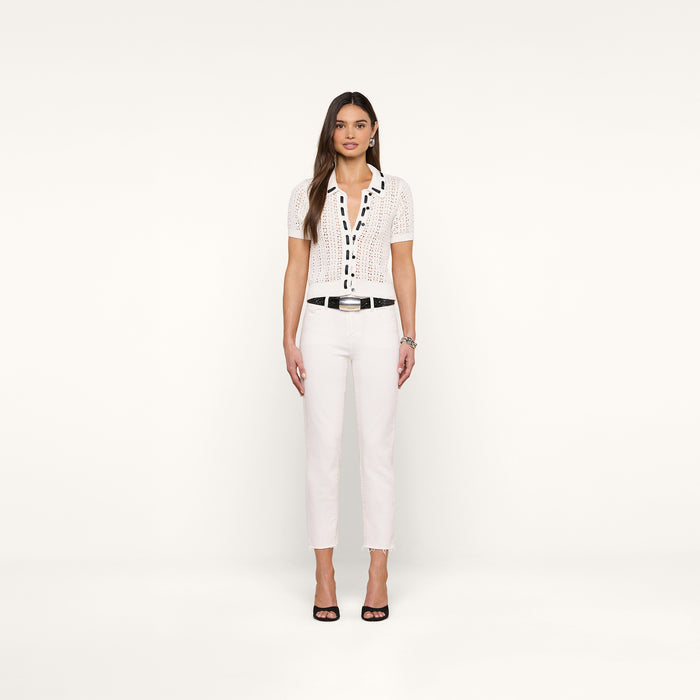 A woman stands facing forward against a plain white background, wearing the Ninette Knit Top—a white short-sleeve crochet top with black accents reminiscent of Parisian style—paired with white pants, a black belt, and black open-toe heels. Her hair is long and loose.