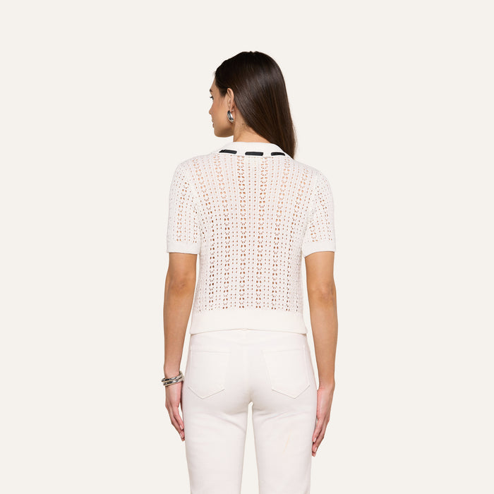 A woman faces away, wearing the Ninette Knit Top—a short-sleeve white top with a Parisian flair and eyelet pattern—paired with white pants. Her long brown hair is down, accessorized with bracelets on her right wrist and large hoop earrings. The background is plain off-white.