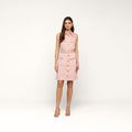 A woman stands facing forward, wearing the Noam Poplin Shirt Dress—a sleeveless light pink cotton poplin piece featuring large front pockets and gold buttons—paired with beige heels, set against a plain white background.