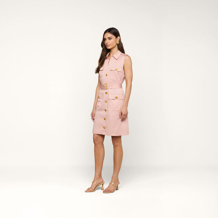 A woman stands and poses against a plain white background, wearing the Noam Poplin Shirt Dress—a light pink, sleeveless dress crafted from cotton poplin with gold buttons, a waist belt, and pockets—paired with nude heeled sandals.