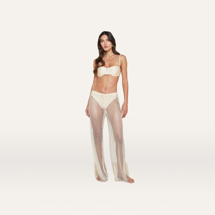 A woman stands on a plain white background wearing a cream bra and the Noemi Mesh Cover-Up Pant in champagne, featuring a flared leg.