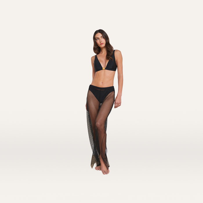 A woman stands against a white background wearing a black bikini top and the Noemi Mesh Cover-Up Pant, barefoot, with one knee slightly bent and her arms relaxed at her sides.
