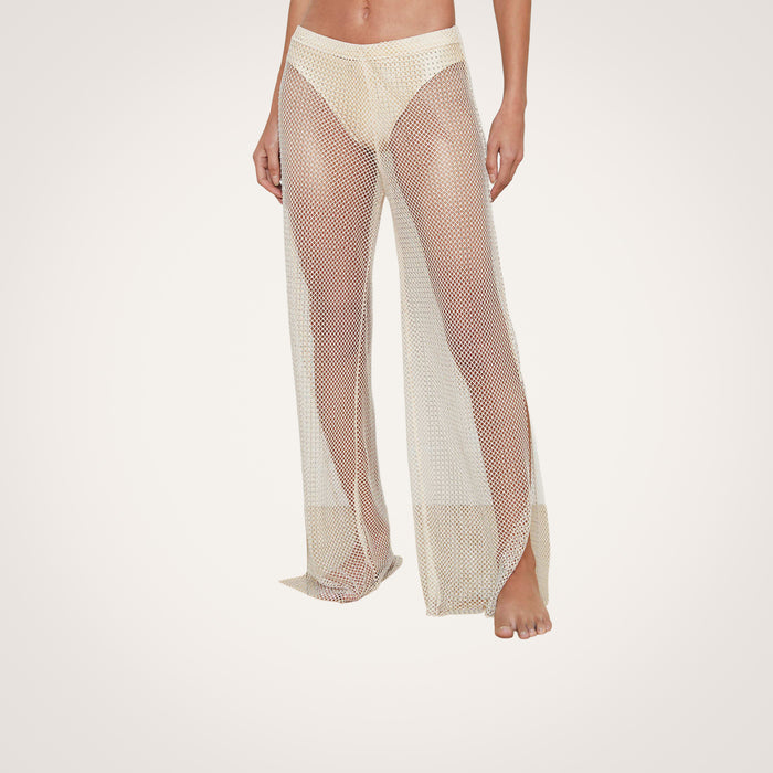 Wearing the Noemi Mesh Cover-Up Pant, a person stands barefoot against a plain light background. The beige pants feature a sheer mesh fabric with flared legs, layered over nude underwear.