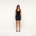 A woman wearing a black tank top, the Norene Nylon Mini Skirt in black with cargo pockets, black heels, and sunglasses stands against a plain white background.