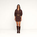 A woman stands against a plain white background wearing the Norene Nylon Mini Skirt—a brown mini skirt featuring a belted design and buttoned pockets—paired with matching knee-high boots, large sunglasses, and gold jewelry.