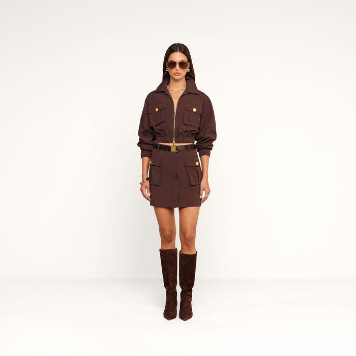 A woman stands against a plain white background wearing the Norene Nylon Mini Skirt—a brown mini skirt featuring a belted design and buttoned pockets—paired with matching knee-high boots, large sunglasses, and gold jewelry.