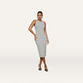 A woman wearing the Nura Racerback Midi Dress—an elegant sleeveless black and white striped jersey design featuring a racerback silhouette—pairs it with black heels and poses with one hand on her hip against a plain light background.