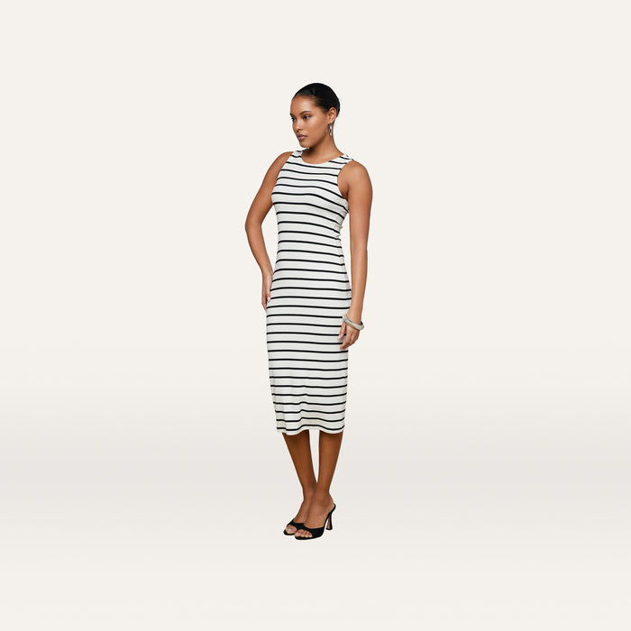 A woman is standing against a plain white background wearing the Nura Racerback Midi Dress—a sleeveless, black and white horizontal striped silhouette—paired with black heeled sandals.