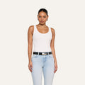 A woman stands against a plain background, wearing the Nyra Bodysuit—a white tank top crafted from soft stretch rib—paired with light blue jeans, a black belt, and hoop earrings. Her hair is pulled back and she has a neutral expression, with her arms relaxed at her sides.