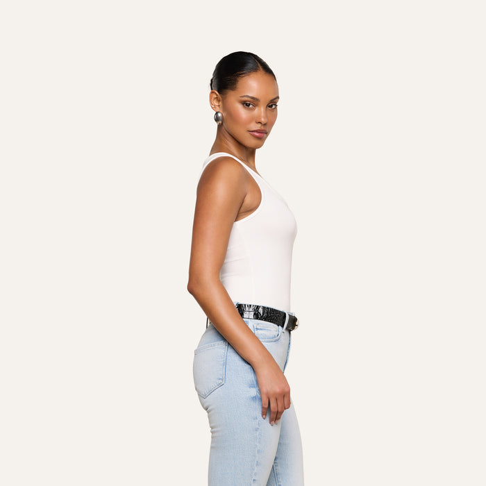 A woman with slicked-back hair wears the white Nyra Bodysuit, light blue jeans, a black belt, and earrings, standing sideways against a plain off-white background.