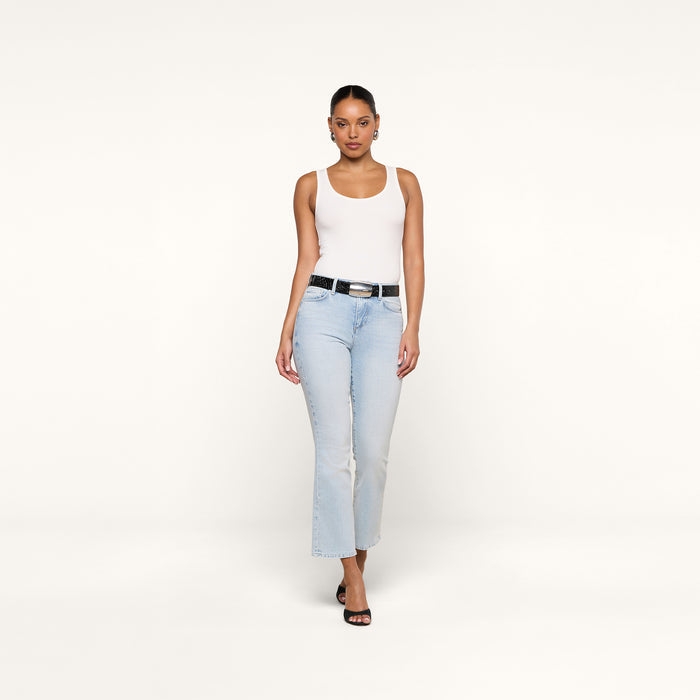 A person wearing the Nyra Bodysuit in white as a base layer, paired with light blue jeans, a black belt, and black heels, stands against a plain white background facing forward with a neutral expression.