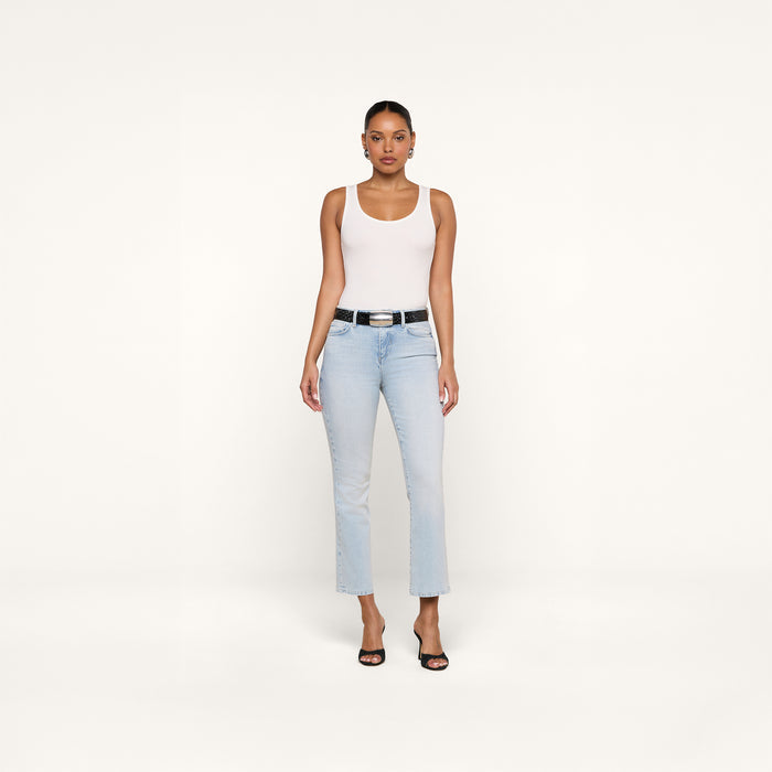 A woman stands facing forward wearing the Nyra Bodysuit as a soft stretch rib base layer, paired with light blue jeans, a black belt, and black open-toe heels against a plain white background.