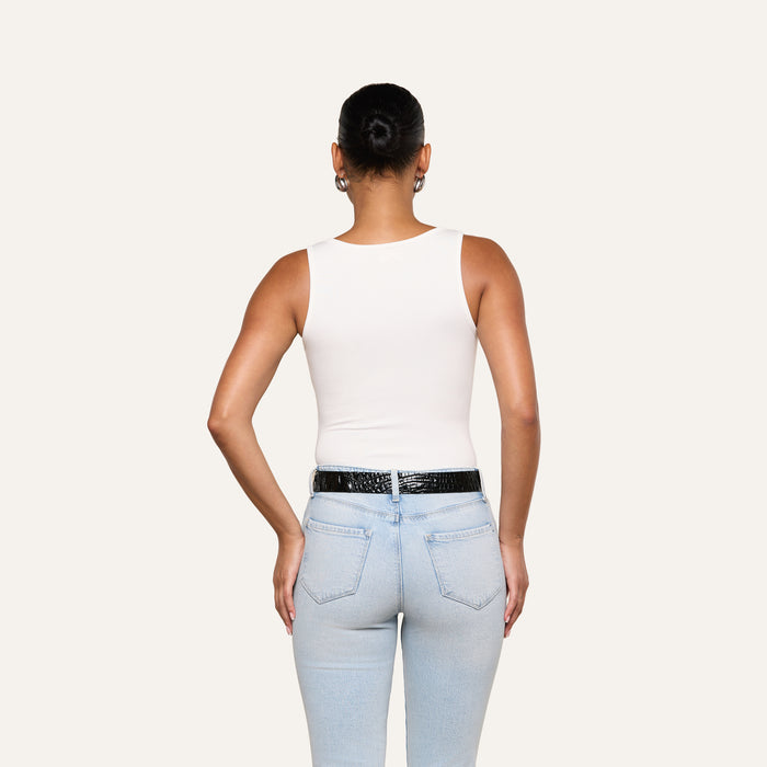 A person with their hair in a bun is seen from the back, wearing the Nyra Bodysuit—a white sleeveless soft stretch rib top—paired with light blue jeans and a black belt, standing against a plain light background.