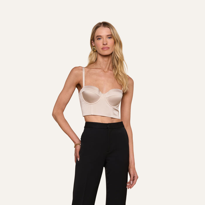A woman wearing the Nysa Bustier Top in light beige—a satin corset-style design with a supportive fit and adjustable straps—paired with black high-waisted pants, stands against a plain white background.