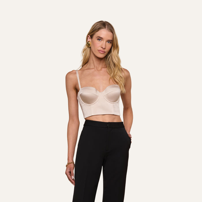 A woman with long blonde hair wears the light beige Nysa Bustier Top with adjustable straps, paired with high-waisted black pants, standing with one hand in her pocket against an off-white background.