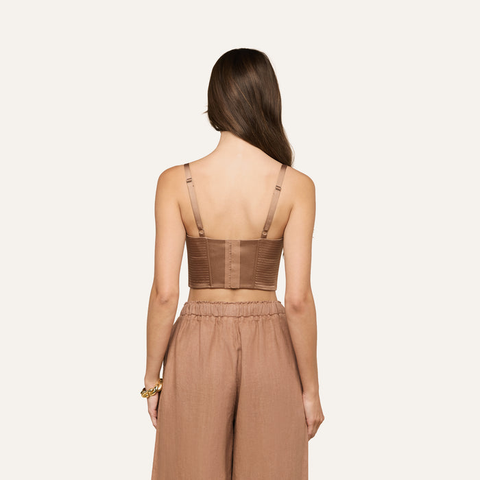 A woman with long brown hair faces away, wearing the Nysa Bustier Top—a brown cropped top with adjustable straps—and loose brown pants. She has a gold bracelet on her left wrist. The background is plain and light.