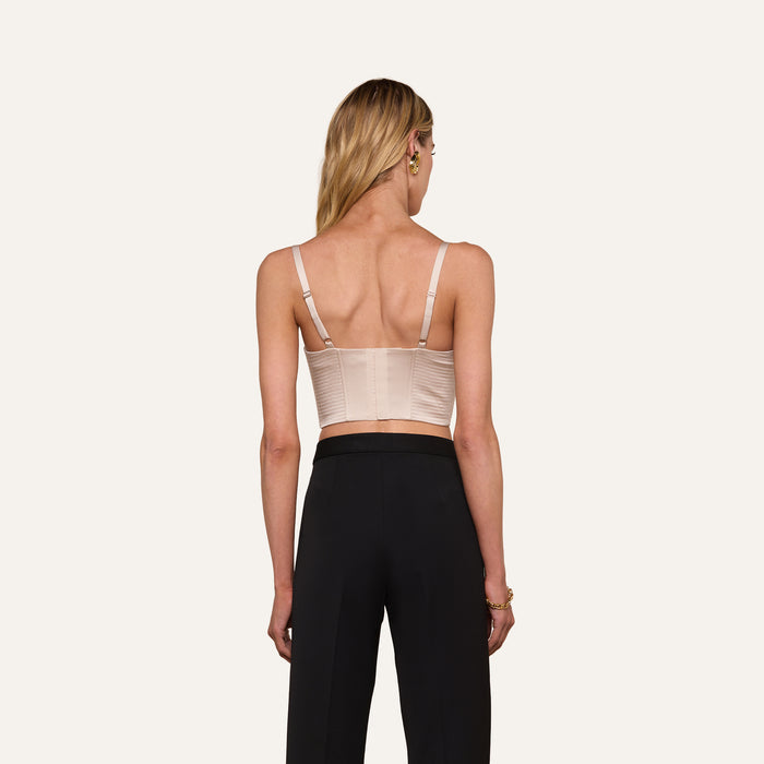 A woman with long blonde hair, seen from behind, wears the Nysa Bustier Top—a beige sleeveless crop top with adjustable straps—paired with high-waisted black pants against a plain background.