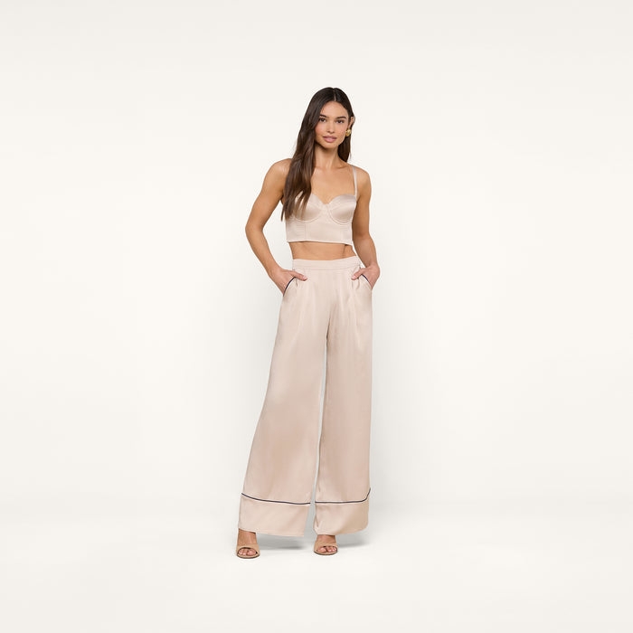 A woman stands against a plain white background wearing the Nysa Bustier Top—a cream-colored crop top styled for a supportive fit—and matching high-waisted wide-leg pants with side pockets and heeled sandals. She has long, loose hair and a neutral expression.
