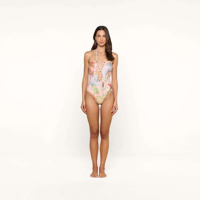 A woman stands against a plain white background, wearing the Opus Halter One-Piece Swimsuit, which features a modern colorful floral and coral reef pattern. She faces forward with her arms relaxed by her sides, showcasing this stylish UPF 50 swimsuit.