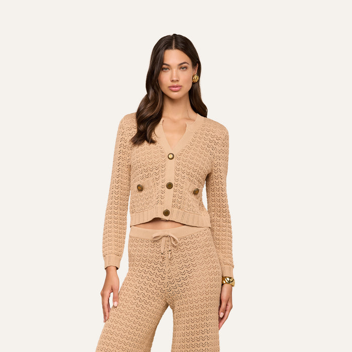 A woman models the Orla Pointelle Knit Cardigan in beige, featuring large buttons and paired with matching knit pants. She stands against a simple light background, her long wavy brown hair styled down and accessorized with gold earrings. Her neutral expression captures an effortless chic resort wear aesthetic.