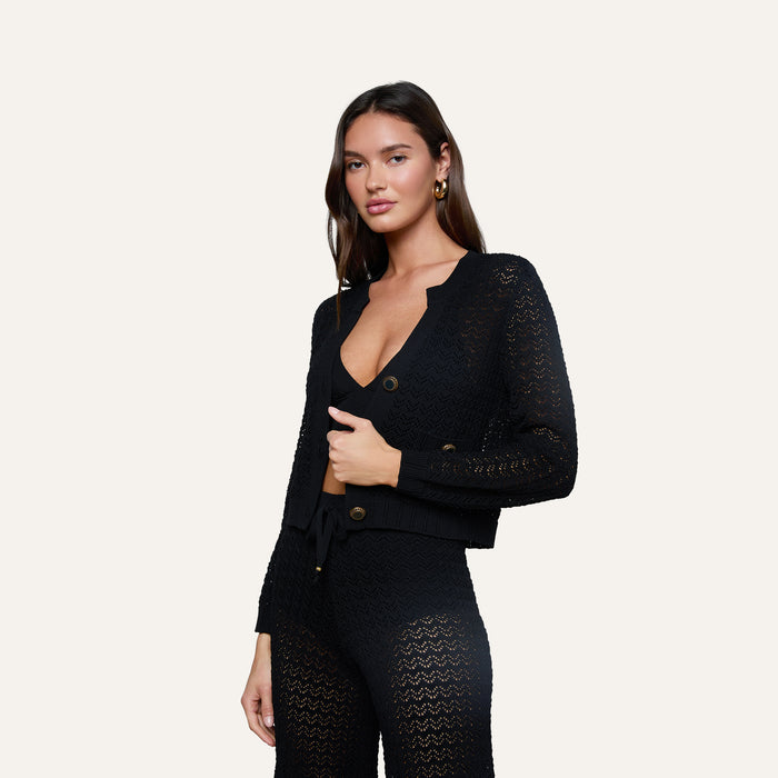 A woman stands against a plain light background wearing the Orla Pointelle Knit Cardigan—a matching black set featuring pointelle knit, a deep V-neckline, and large buttons, paired with coordinating pants.