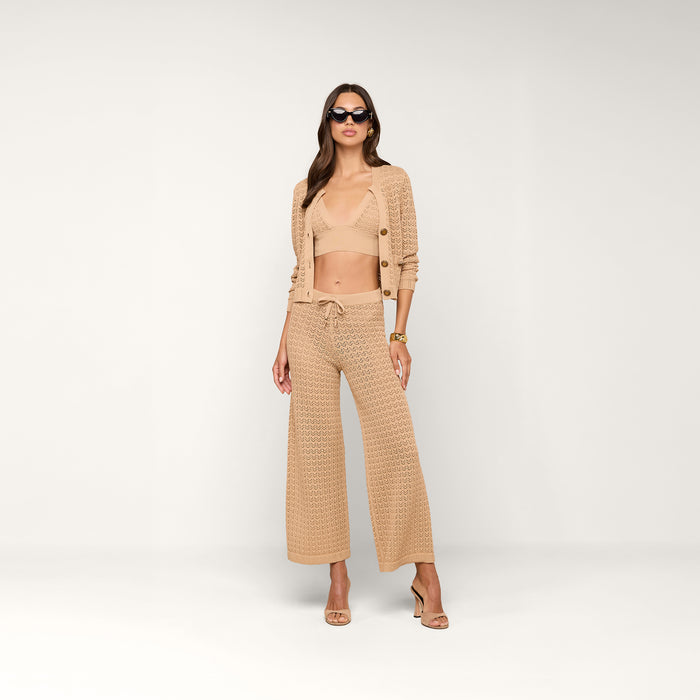 A woman stands confidently in heels against a plain, light background, wearing sunglasses and the Orla Pointelle Knit Cardigan as a lightweight beach coverup, paired with a matching bralette and wide-leg pants.
