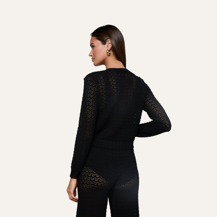 Woman standing with her back to the camera, wearing the Orla Pointelle Knit Cardigan in black, styled with matching pants. She has long brown hair, gold hoop earrings, and stands against an off-white background.