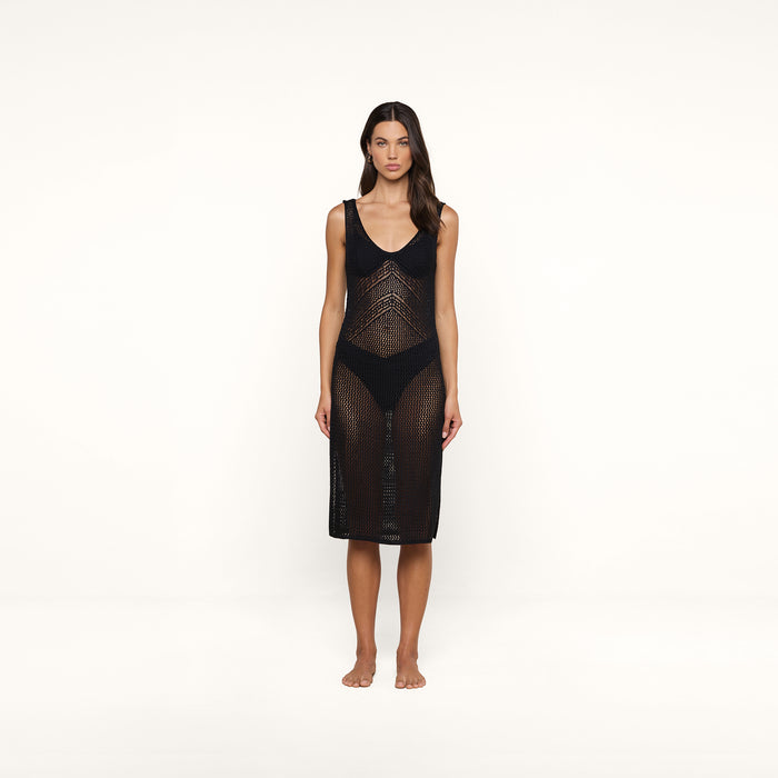 A woman stands barefoot against a plain white background, facing forward with a neutral expression. She wears the Pallas Crochet Cover-Up Dress, a sleeveless, knee-length black mesh design styled over a black bikini.