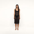 A woman stands barefoot against a plain white background, facing forward with a neutral expression. She wears the Pallas Crochet Cover-Up Dress, a sleeveless, knee-length black mesh design styled over a black bikini.