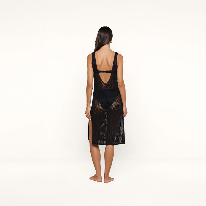 A woman with long dark hair stands barefoot, facing away, wearing the Pallas Crochet Cover-Up Dress—a sleeveless black mesh design with an open back and black undergarments—posed against a plain white background.