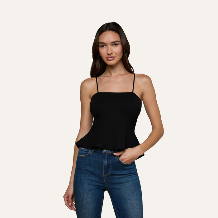 A woman with long brown hair is wearing the Parisa Peplum Top, a black tank top with a fitted bodice, paired with blue jeans. She stands against an off-white background, smiling softly with one hand relaxed by her side.